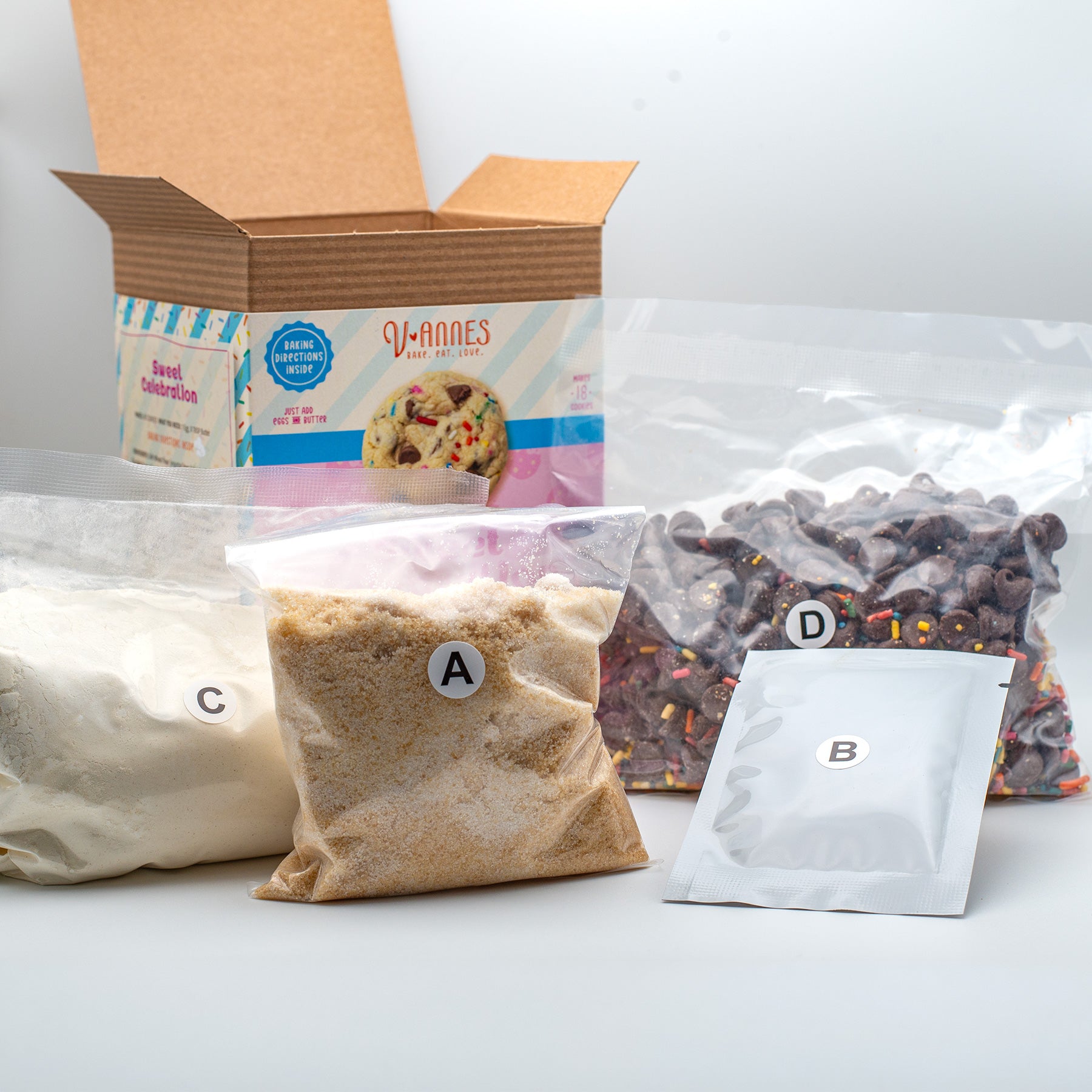 An open cardboard box labeled V-Annes Sweet Celebration shown with four labeled ingredient pouches on a light gray studio background.
